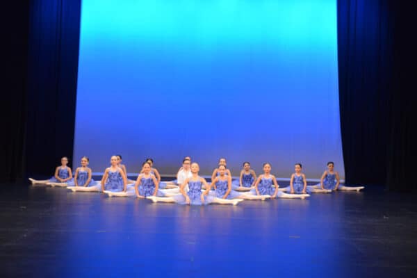 Junior dancers in lilac dresses seated in a line on stage